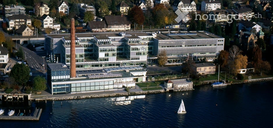 Modern building complex with several floors, glass facade, large parking area, waterfront location, surrounded by trees and other buildings.