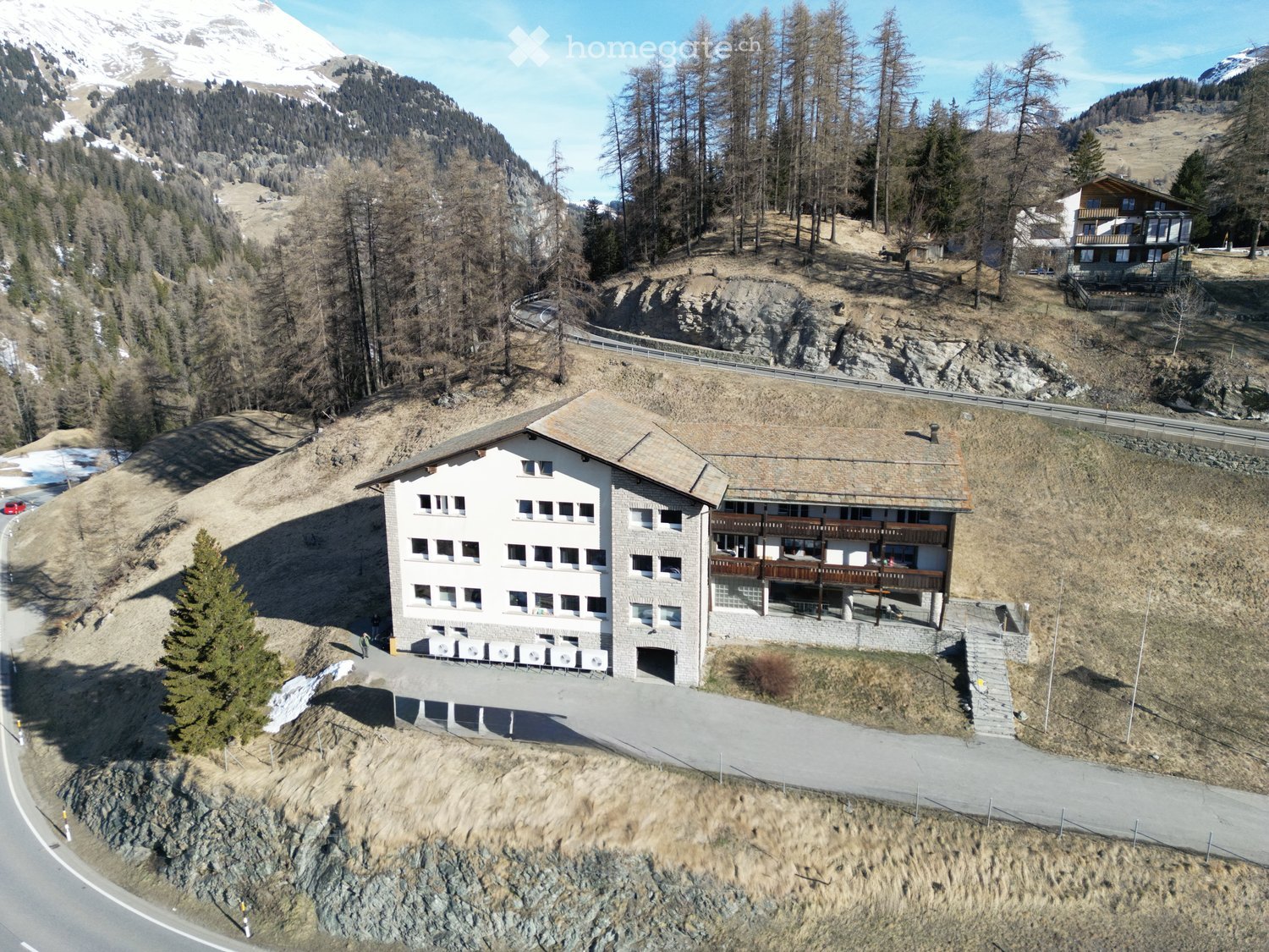 3 story building, white walls, brown roof, balcony, surrounded by trees, mountains in the background