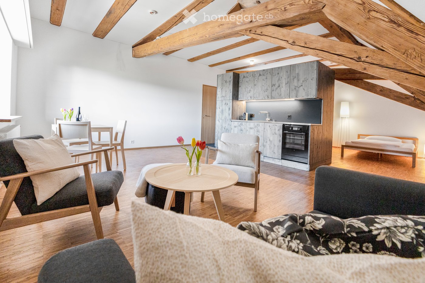 A modern attic room featuring a kitchen with oven, a living area with couches, a dining table with chairs, and a bed in the corner.