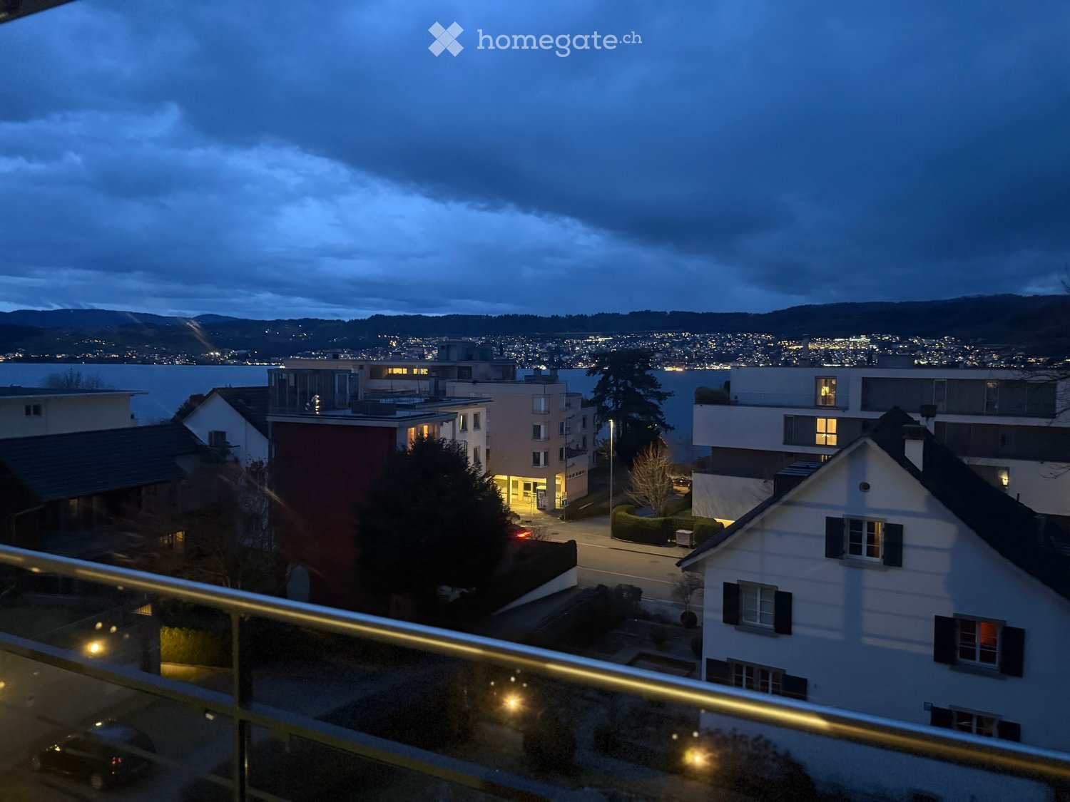 A modern house from a balcony with a glass railing, overlooking a street and cityscape with a lake in the distance, at night.