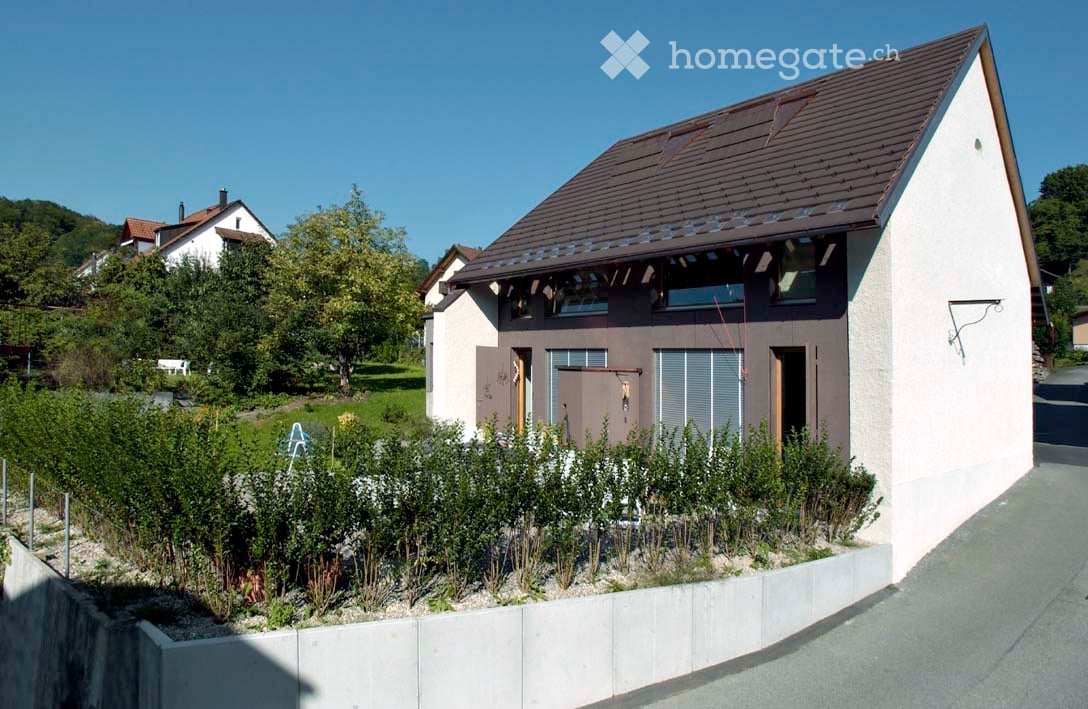 2-story modern house, white and brown walls, brown roof, entrance with garage door, small garden with hedges in front