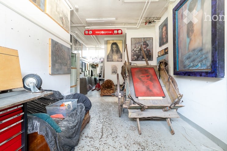 Art studio with multiple artworks on the wall, storage boxes, canvas covered objects, tables, trolleys, chairs