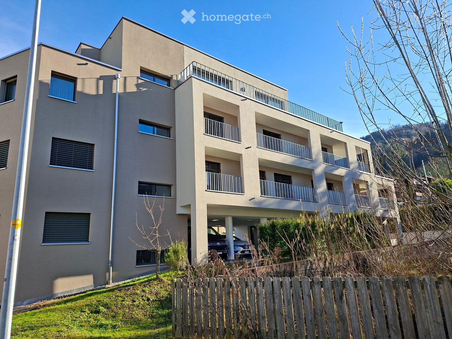 Modern multi-story building, beige exterior, multiple balconies, attached garage, fenced yard, lush greenery, clear sky