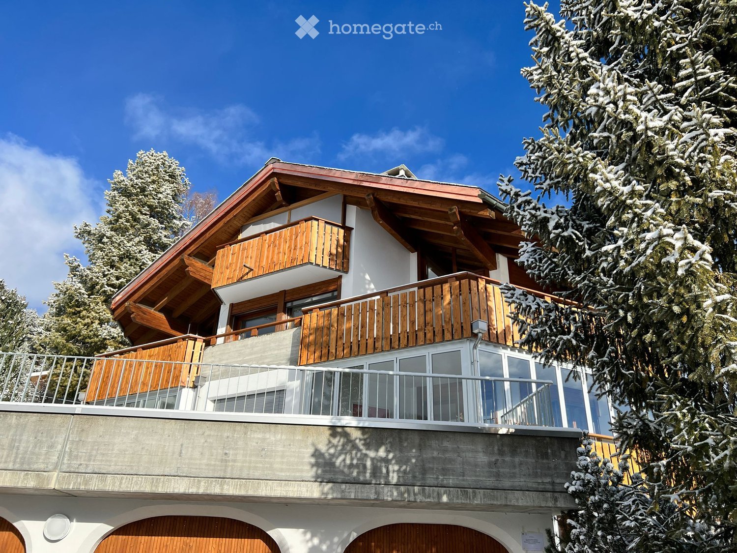 2 story chalet, red roof, wooden balcony, concrete raised ground floor, wooden garage doors