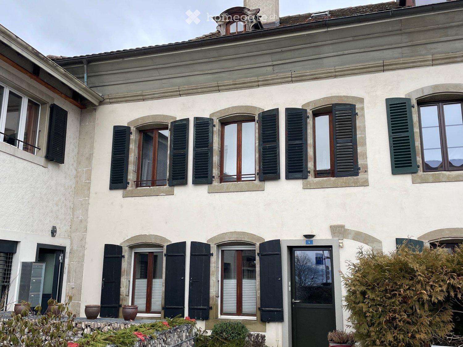 Large building, white walls, black shutters, brown windows, large entrance door, garden with potted plants, bushes, flowers