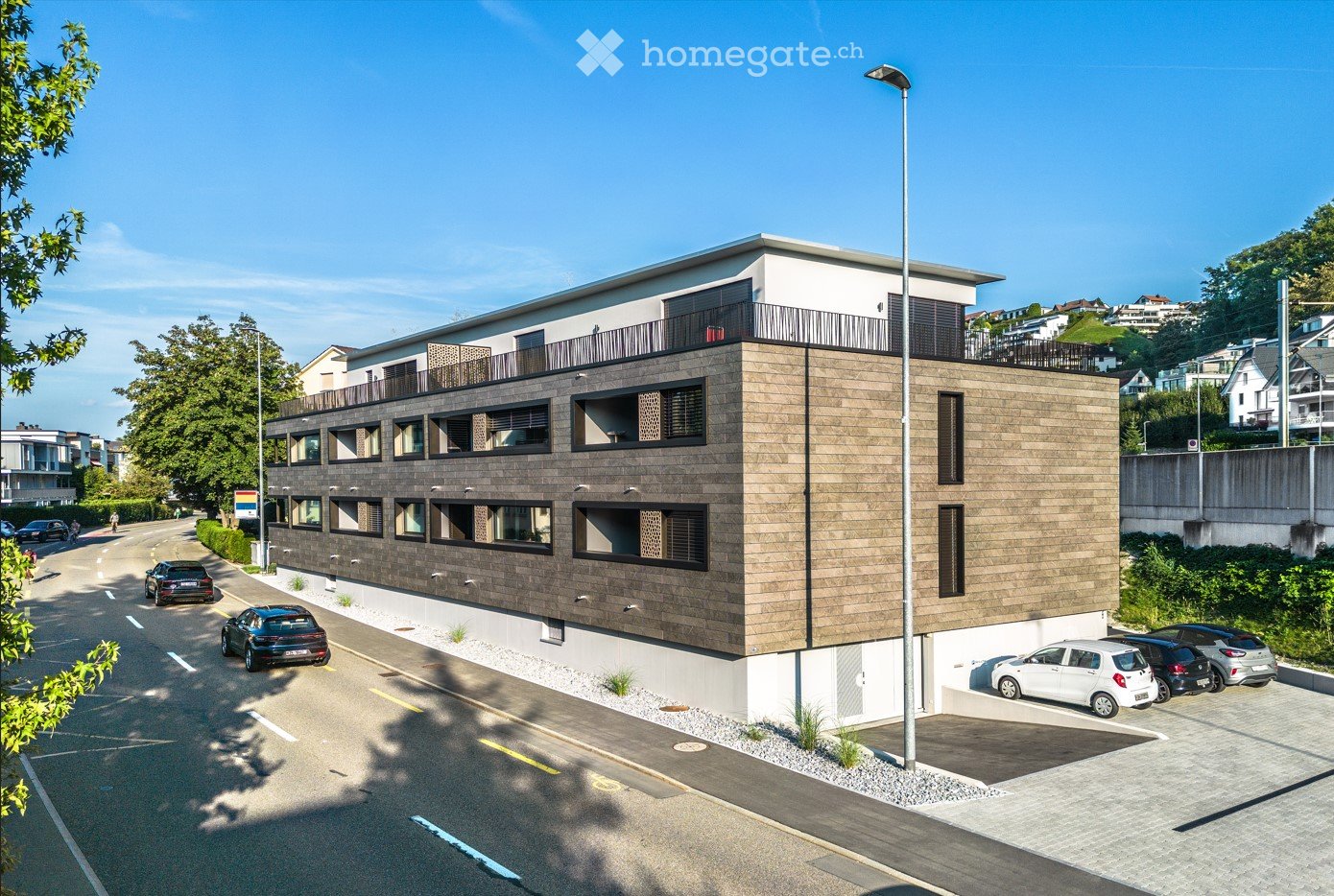 Multistory apartment building with brown stone wall and white roof, balconies, parking space for cars.