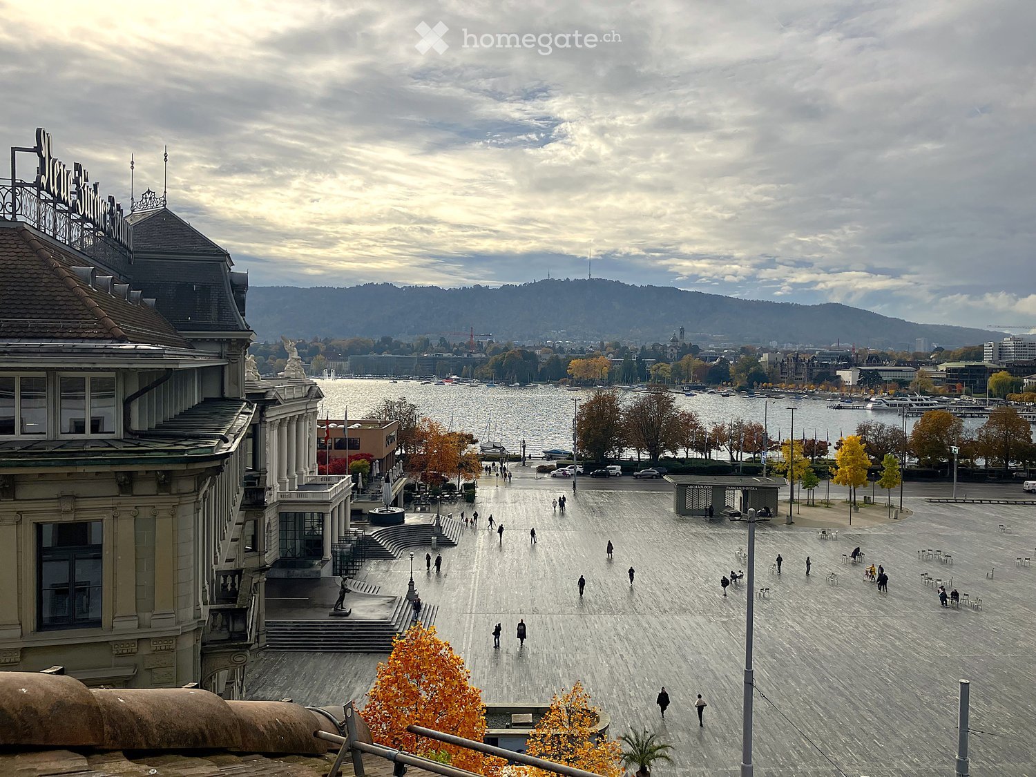 Outdoor square with people walking, buildings, lake, mountains in the distance