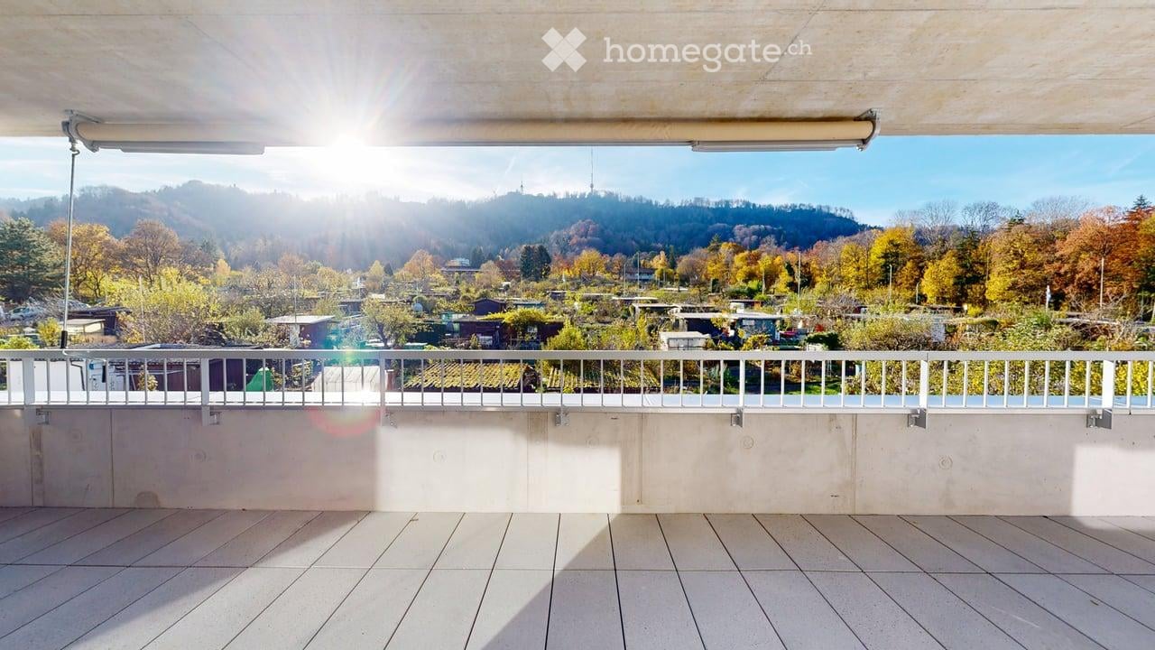 A balcony with white tiled flooring and a white railing, offering a view of a garden and mountainous area