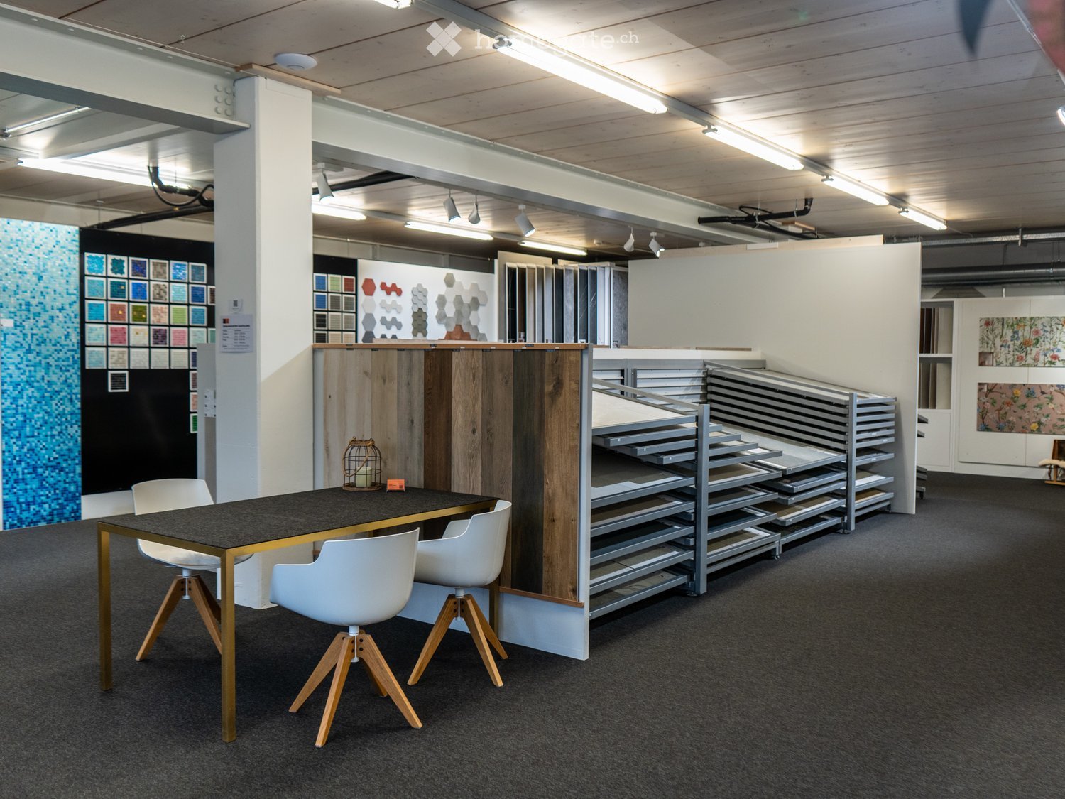 Empty modern showroom with carpet flooring, steel racks filled with building materials, dining table with chairs, wall display with colored tiles
