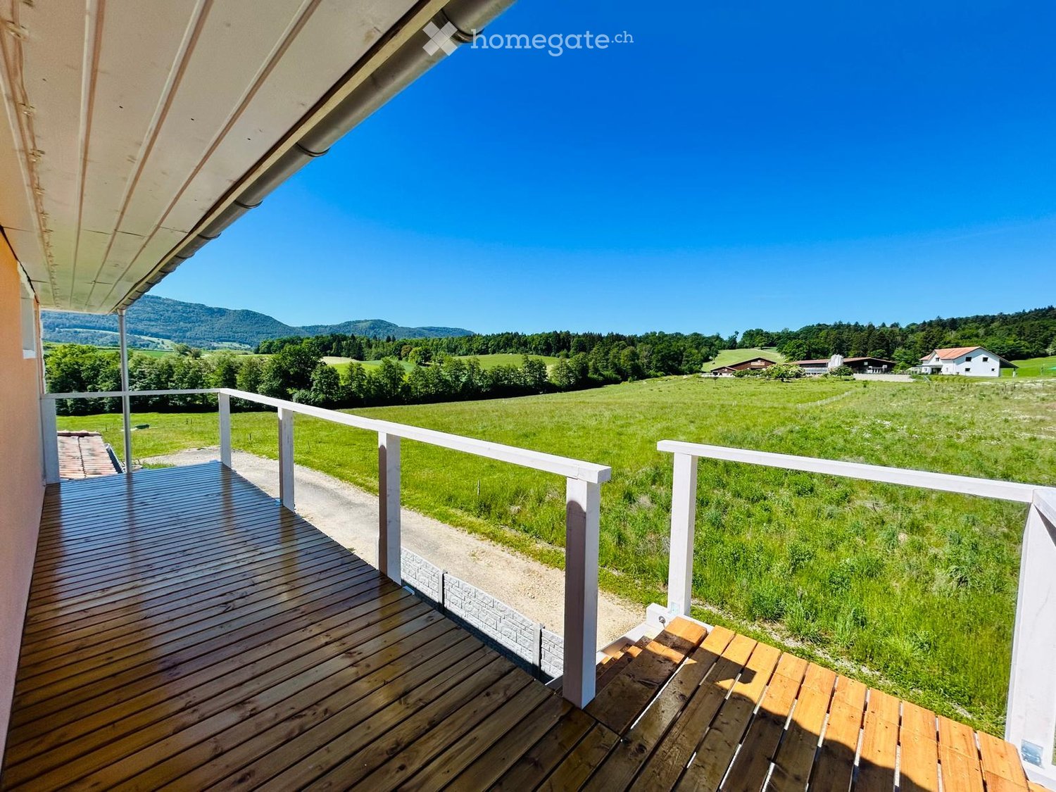 Wooden balcony with white railing, overlooking a green field and distant mountains under a clear blue sky.