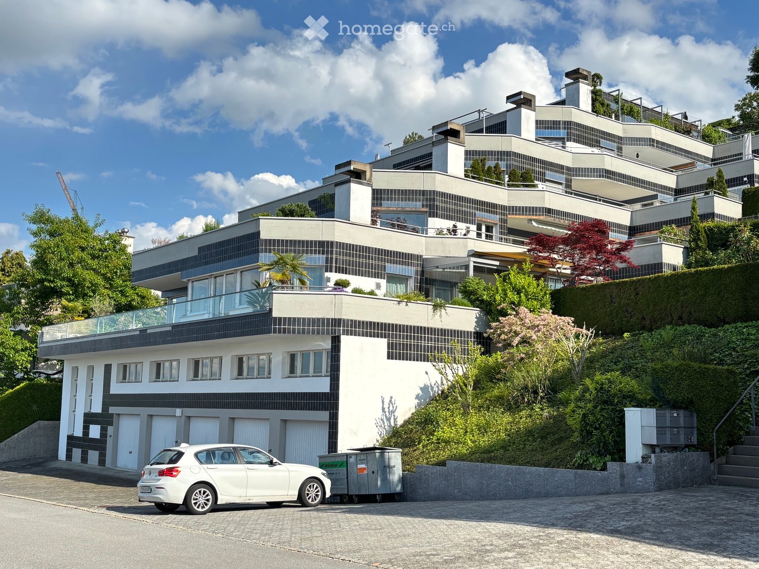 Modern multi-story house with white and black facade, large windows, balconies, and a garage. Surrounded by greenery and a white car parked in front.
