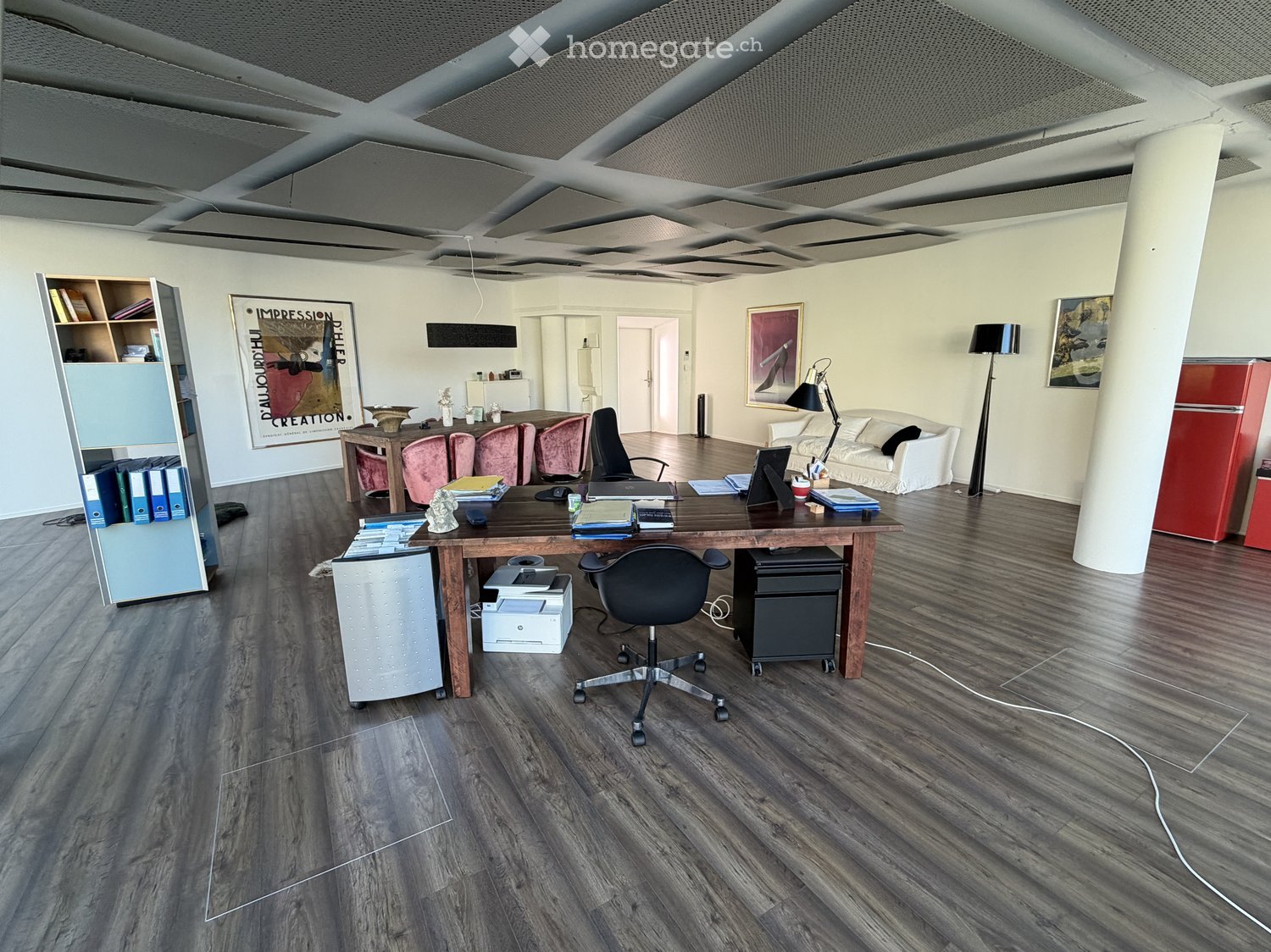 A spacious room with large wooden floor, wooden desk with chair, printer, books, dining chairs, tables, artwork, lamps, and a white sofa.
