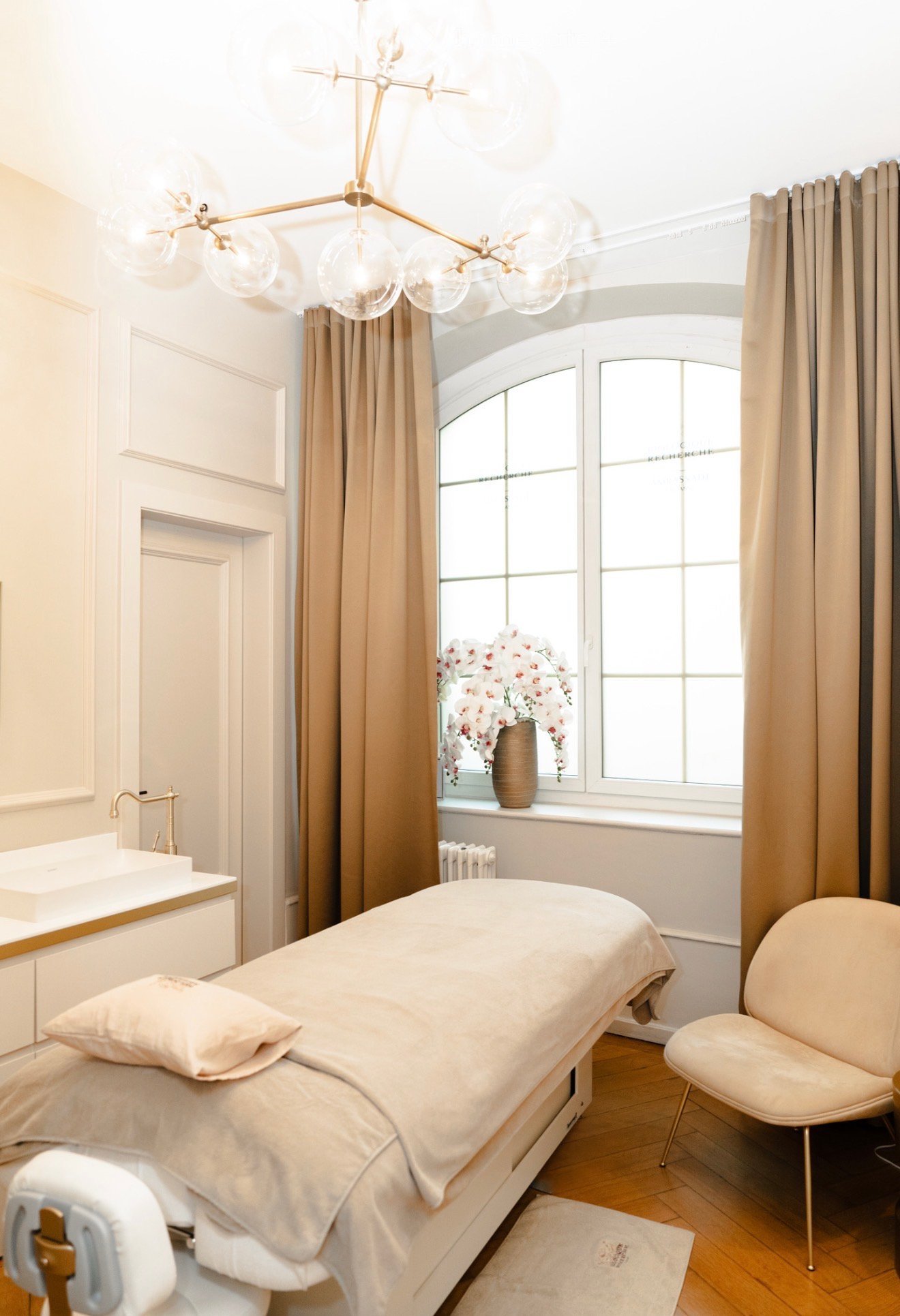 A spa room with a bed, a sink, a faucet, a chair, a chandelier, a window, and a flower vase