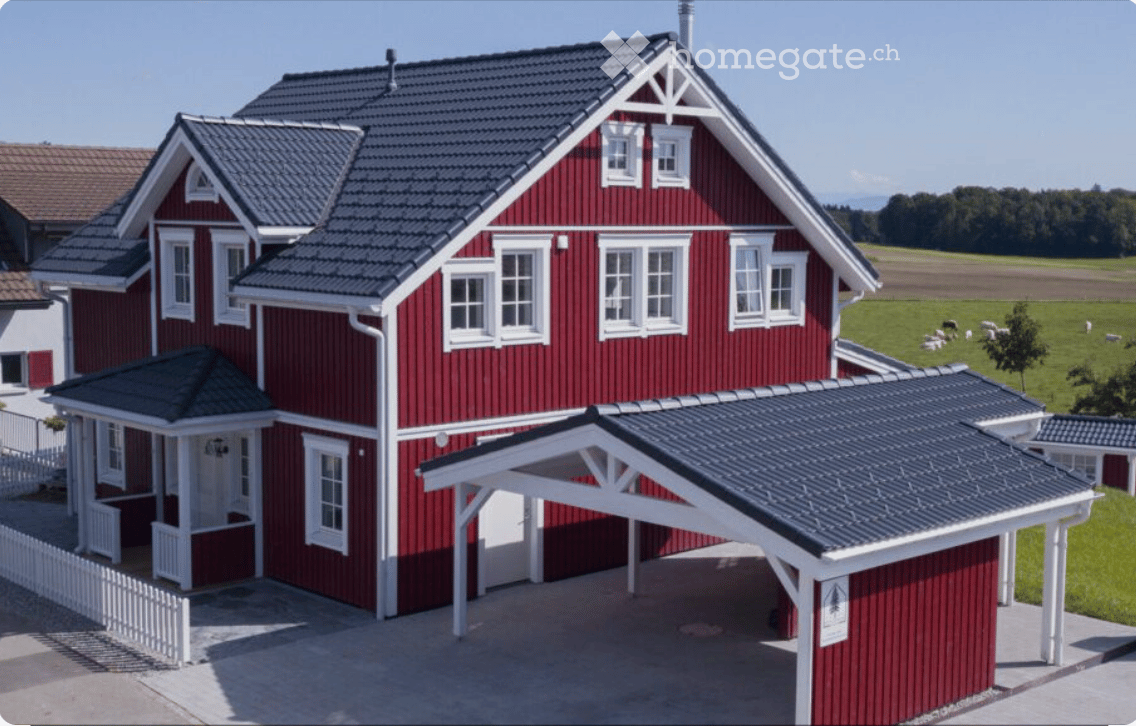 House. Red wooden house with white trim, multiple windows, and a covered driveway. The house features a pitched roof and is surrounded by a green field with grazing animals.