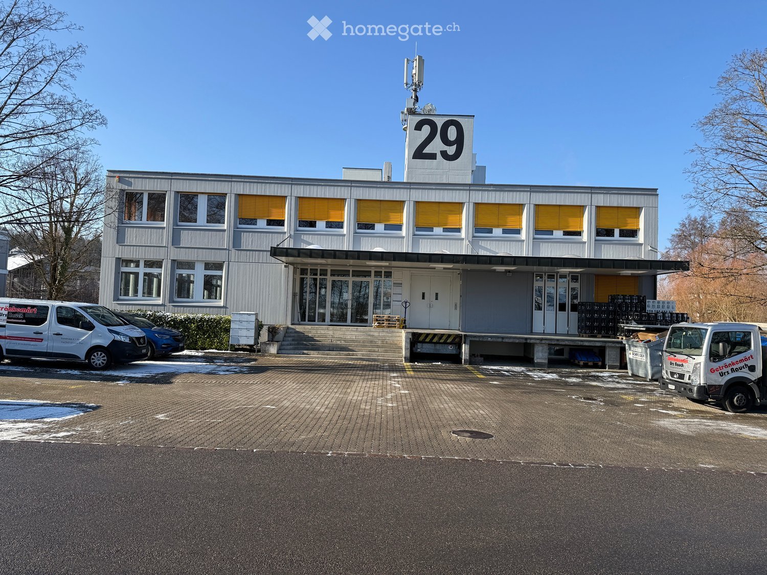 Large building with multiple windows, number 29 on the top, entrance with stairs, parking lot with vans, bushes, snow