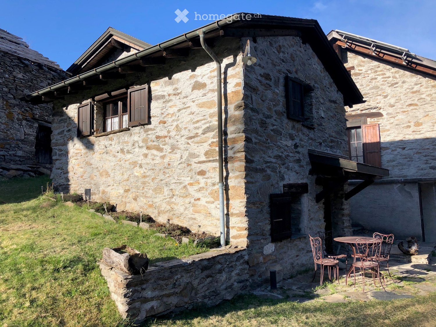 Stone wall house with wooden windows and doors, outdoor table with chairs, located on a sloped grassy terrain