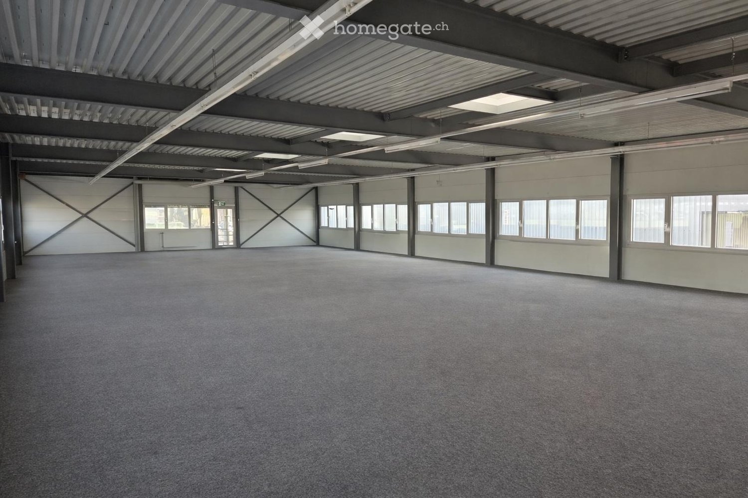 Empty warehouse with gray flooring, metal beams, and large windows.