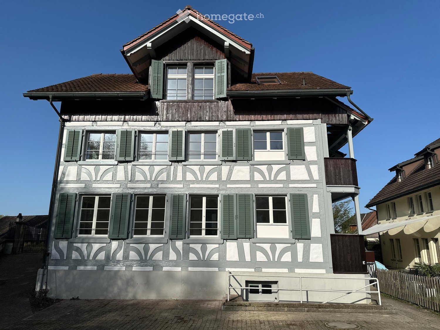 a three story house with wooden trim and shutters, many windows, brown roof, white and brown walls, balcony, fenced garden in front