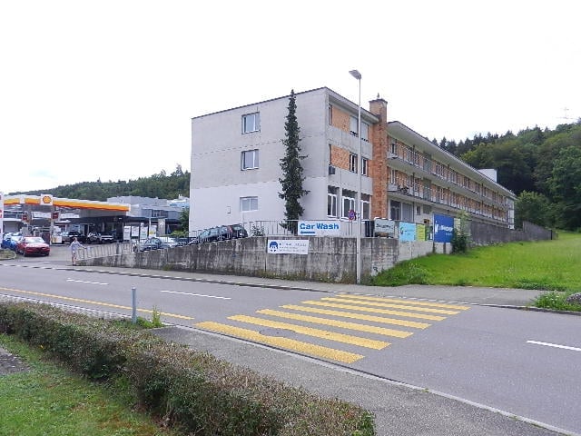 Multistory building, several windows, car wash sign, cars parked on the street, trees, streetlight, gas station