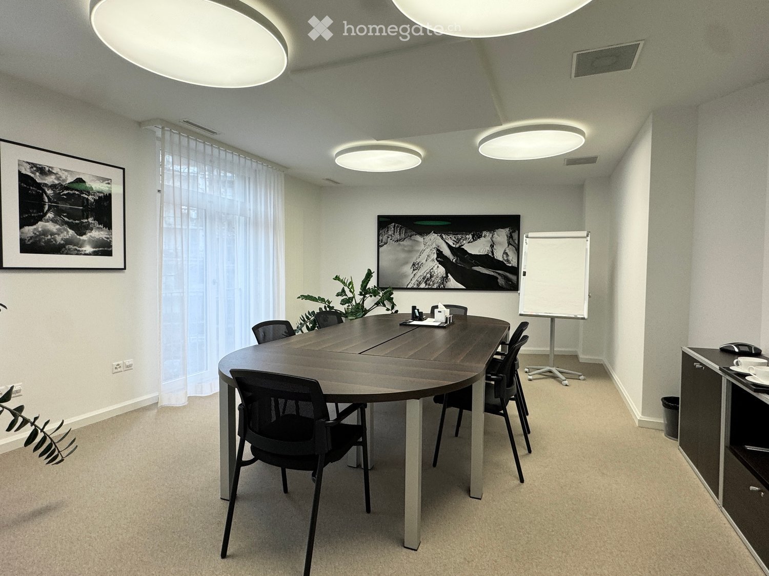 An empty conference room, oval-shaped wooden desk, dark-colored chairs, mounted picture frames, whiteboard, trash bin, and wooden shelves with electronic equipment.