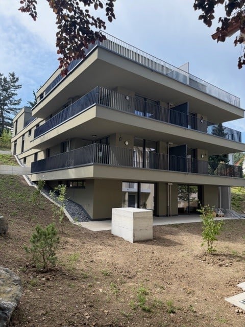 Modern multi-story residential building with multiple balconies and raised ground floor, surrounded by greenery.