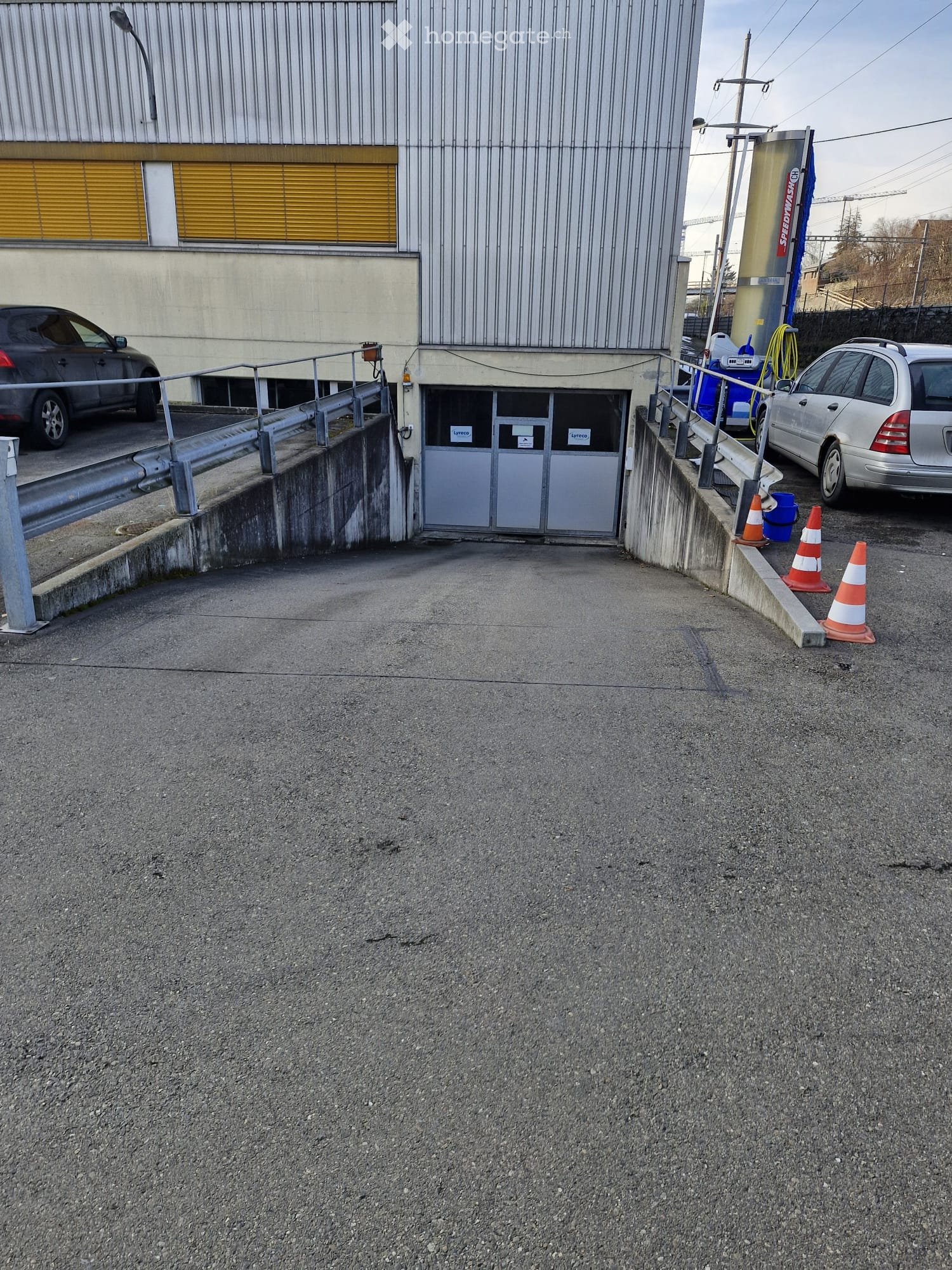 Commercial building with underground garage access, metal railings, grey parking area, orange traffic cones, parked cars, yellow shutters, white and yellow stripes on building