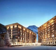 Modern hotel complex with multiple buildings, large windows, ground lighting, snowy landscape, mountain range in the background