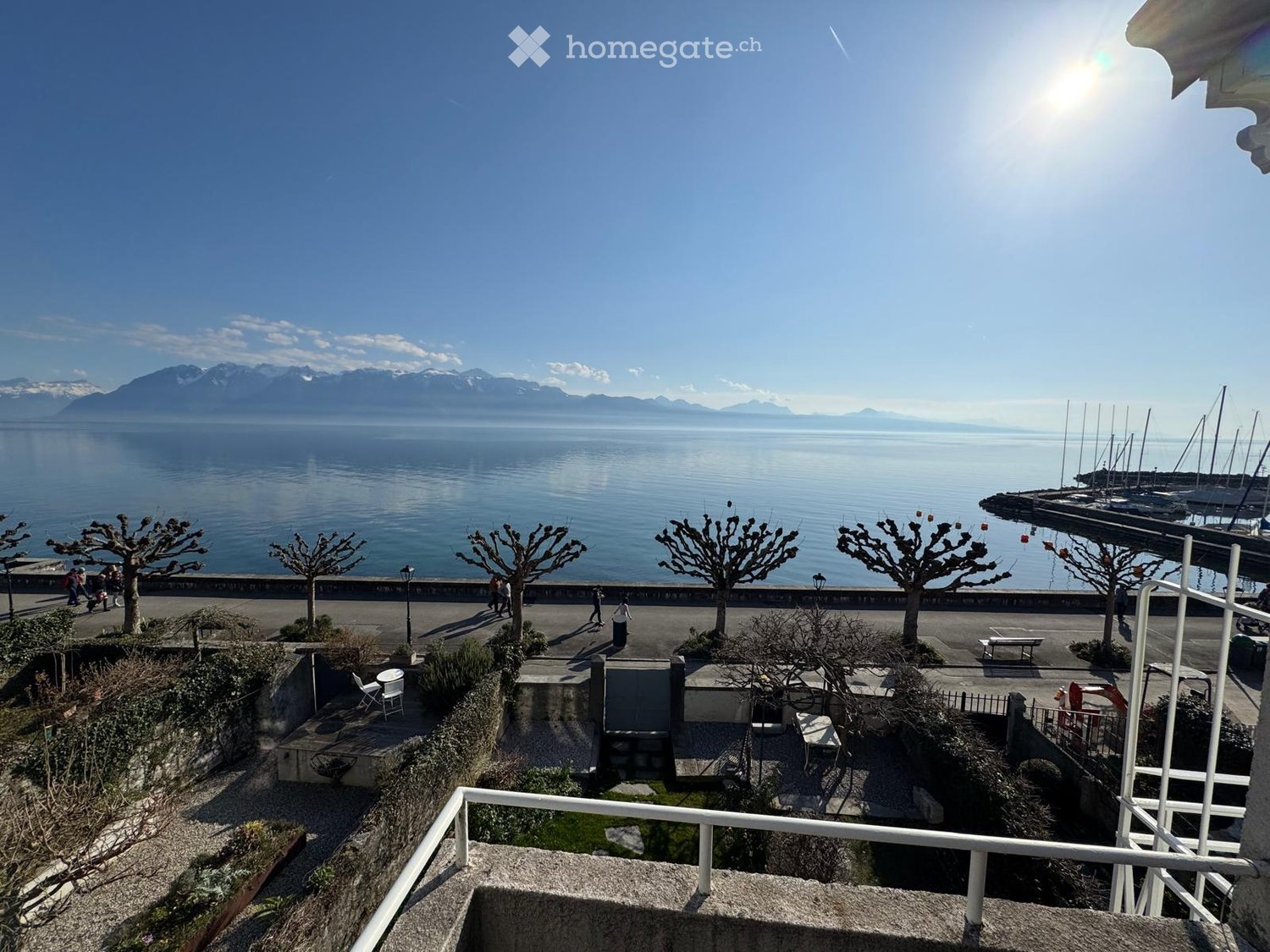 Waterfront promenade with people walking, row of trees, lake view with mountains in the distance, clear blue sky
