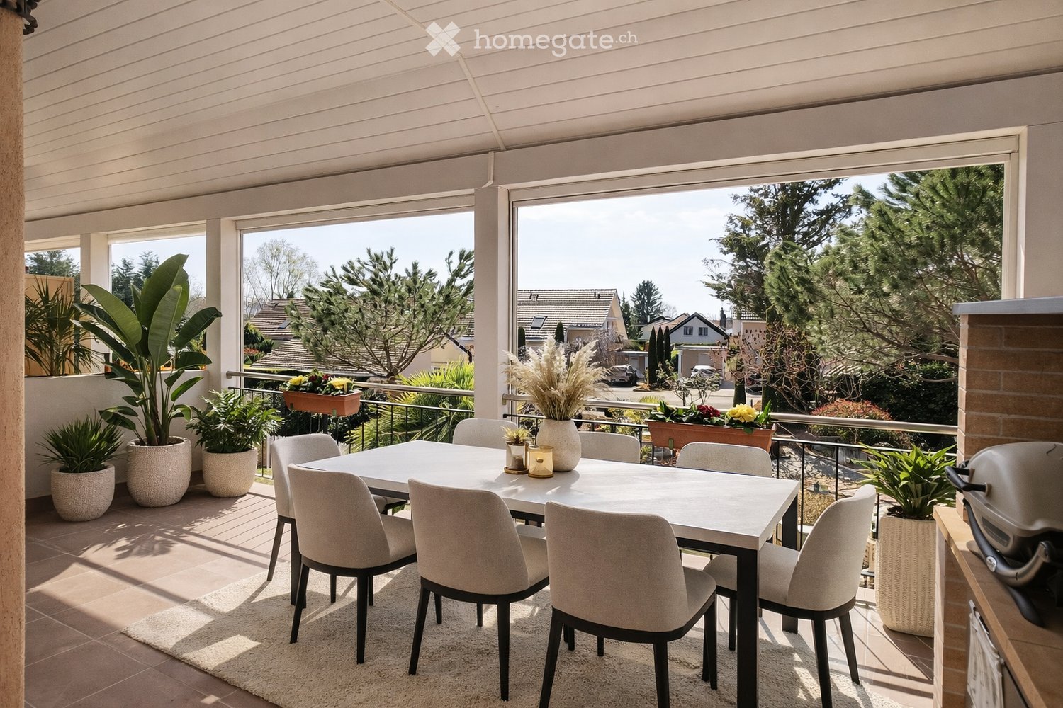 Balcony / Terrace. Balcony with a modern dining setup, including a white table, six chairs, a vase with dried flowers, and several potted plants. The balcony has a view of a suburban neighborhood with houses and trees.