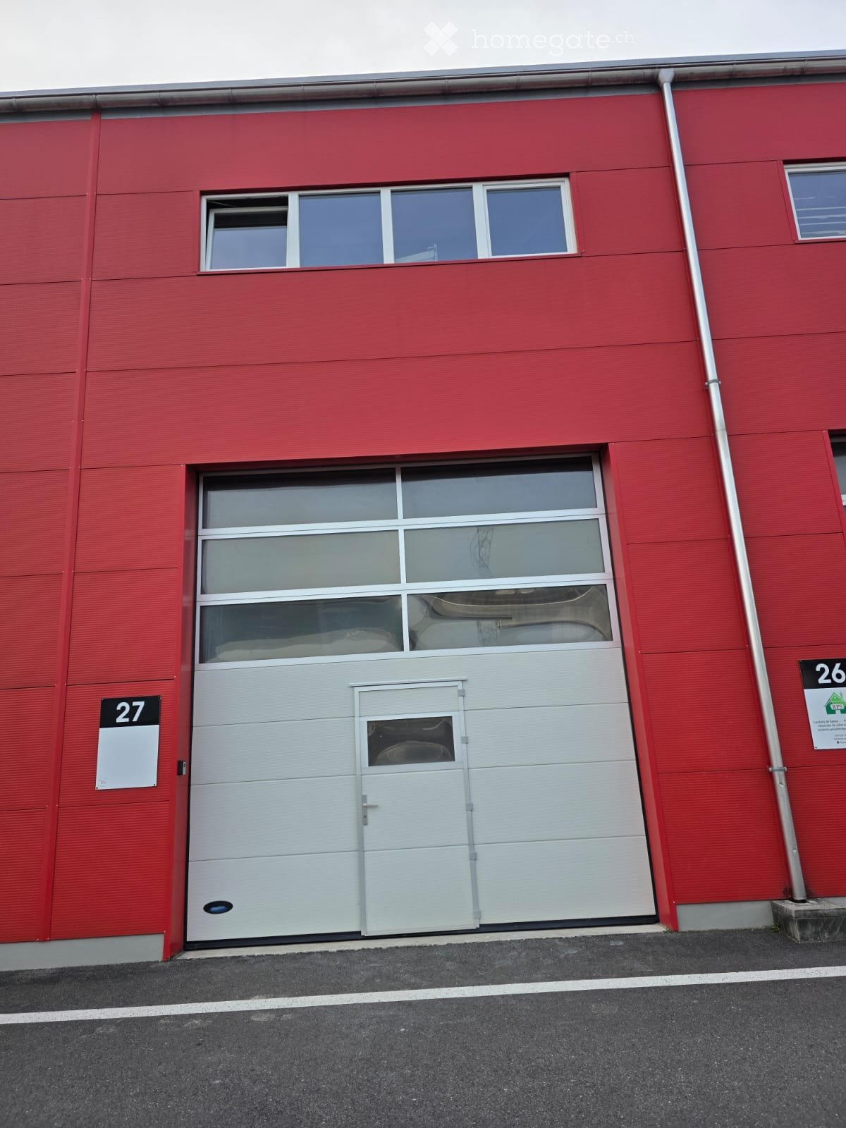 Red building with a single garage with double doors, two small windows above the door and a number board with the number 27