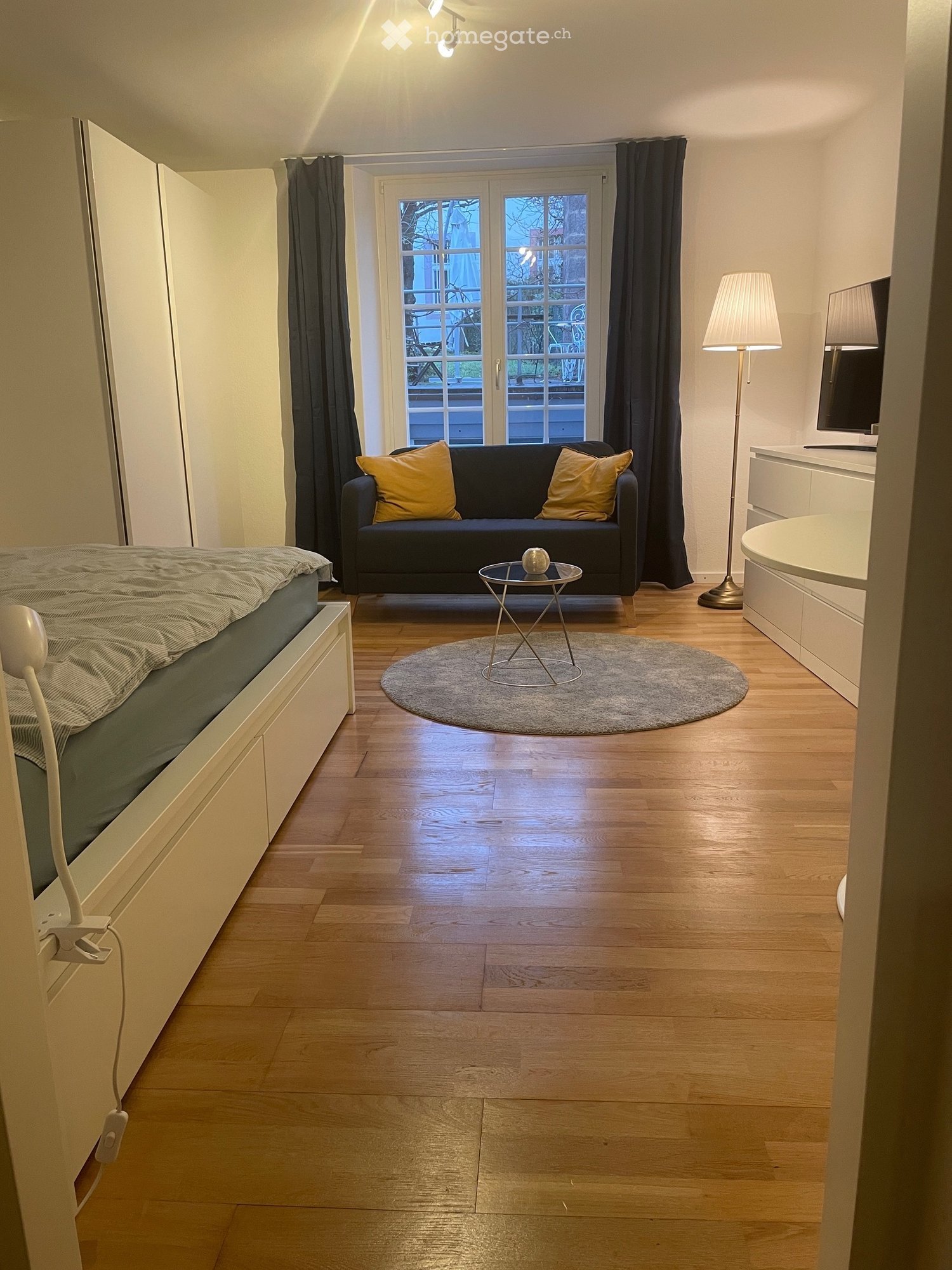Modern interior with a white bed, a grey couch with yellow cushions, a small table, and large window