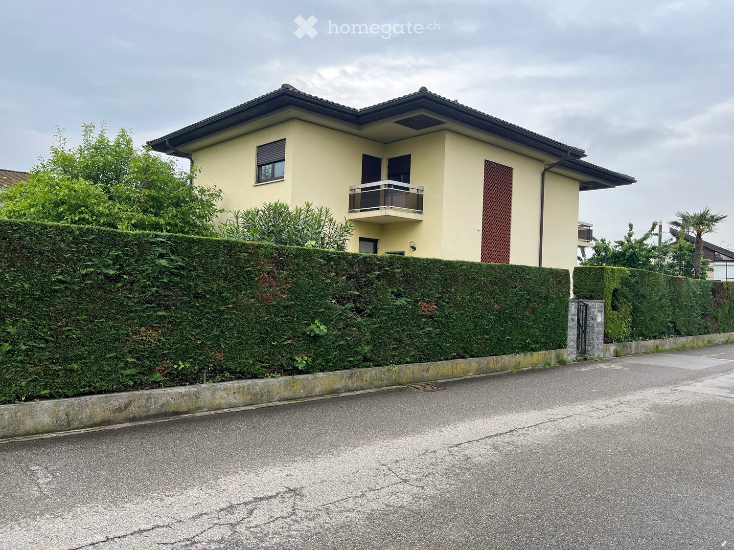 two-story house, yellow exterior, black roof, window with shutters, balcony, hedge in front, brick sidewalk