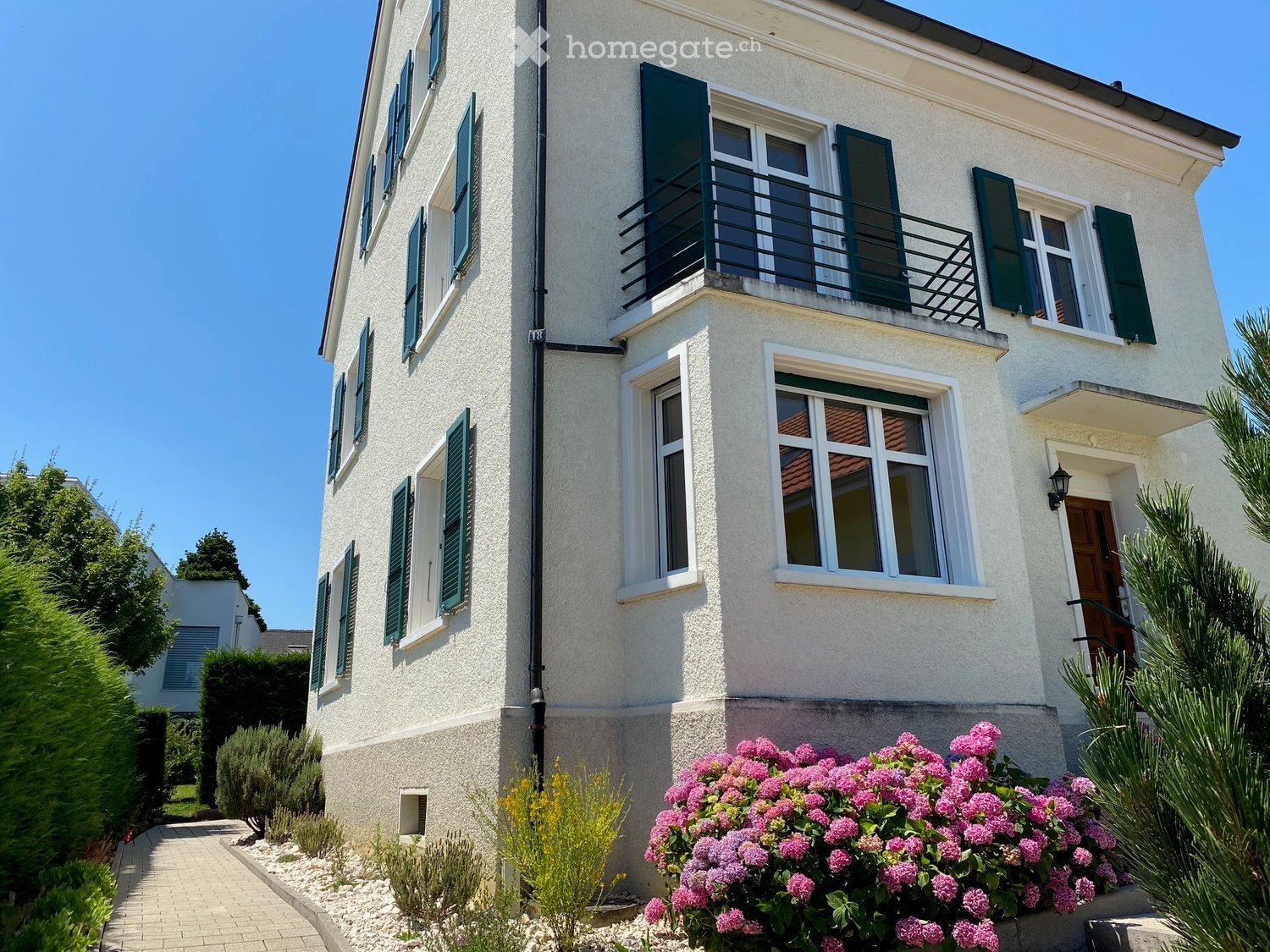 Three-story house, green shutters, white exterior, paved entrance, black balcony railing, pink flowers