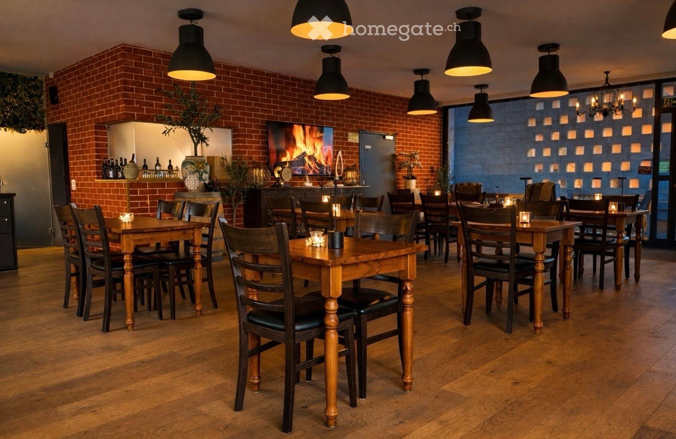 Dining area with brick walls, tables, chairs, lit candles, wooden flooring, fireplace, chandelier, shelves with bottles and vases