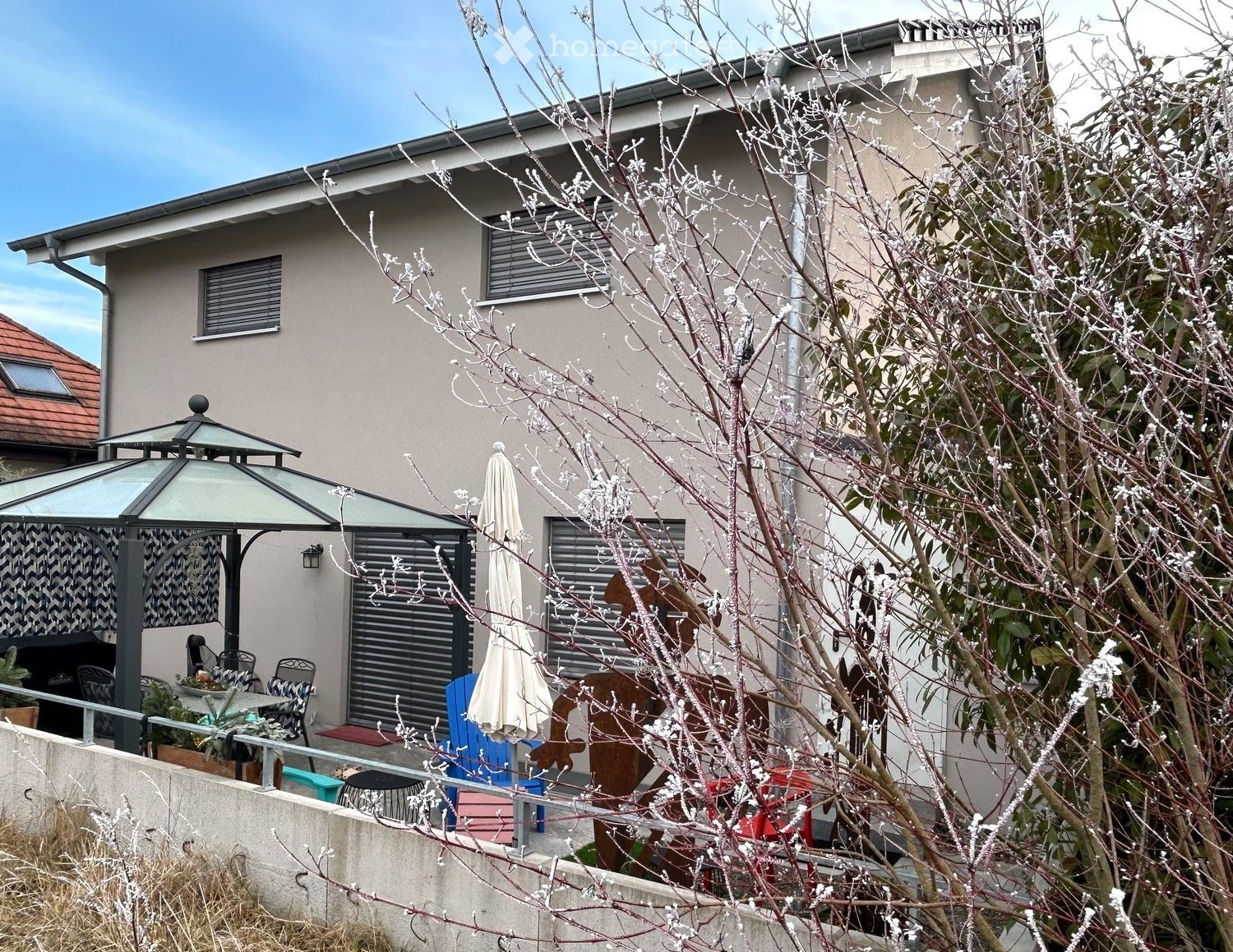 Modern residential house with terrace and garden, located at the edge of a village, featuring white and gray color scheme, glass and metal gazebo, outdoor furniture, and a metal sculpture.