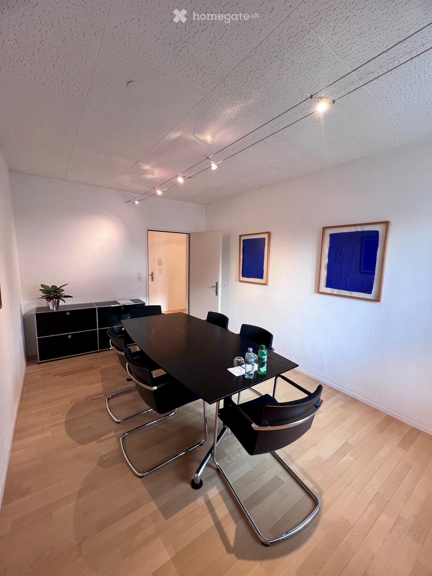 A room with a conference table, office chairs, framed artwork on the wall, potted plant on a cabinet, white walls, and wooden floor.