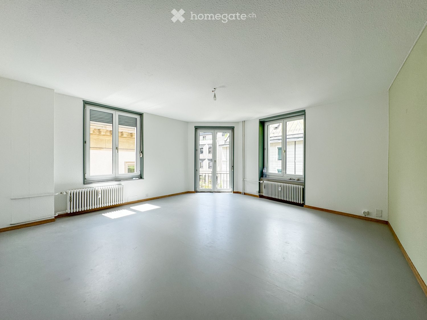 Empty room with light grey walls, white floor, and large windows with metal frames. Radiators are visible beneath the windows, and the space is completely unfurnished.