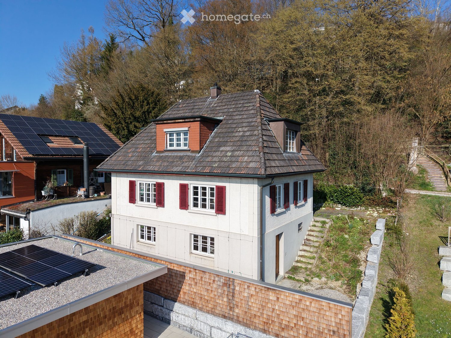 2 story house, white exterior, red shutters, solar panels on roof, steps leading to entrance, surrounded by trees