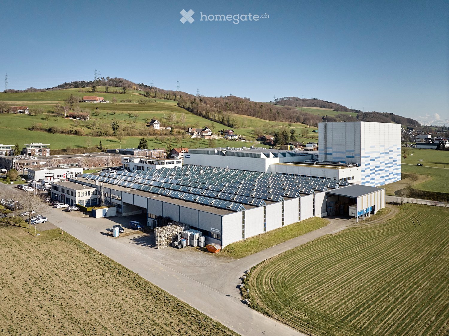 Large industrial building with solar panels on the roof, surrounded by fields and a parking lot