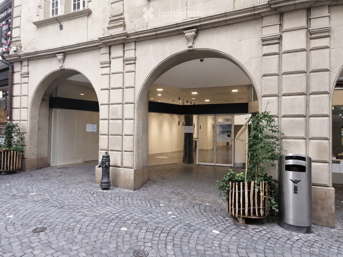 Entrance to a building with arched doorways, potted plants, and a trash bin on a cobblestone area.