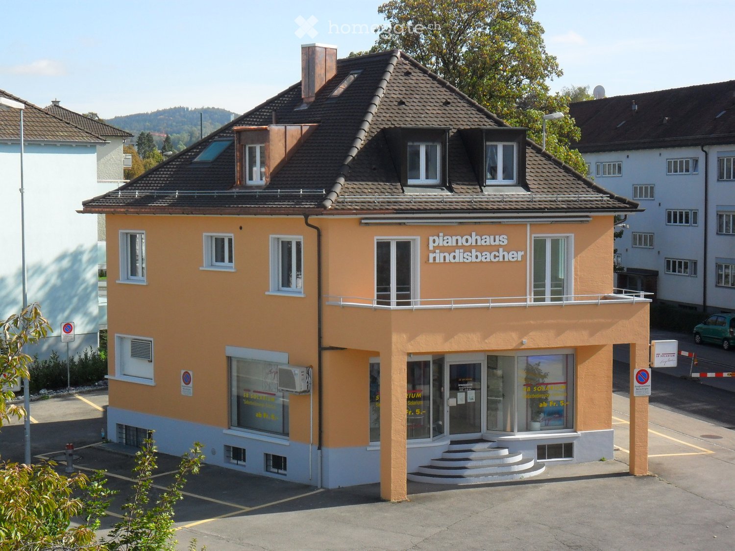 Two-story building with a brown roof, yellow walls, white windows, and a balcony on the second floor. The building has a sign that reads 'pianohaus rindisbacher'. There is a parking lot in front of the building, and a green car is parked on the side of th