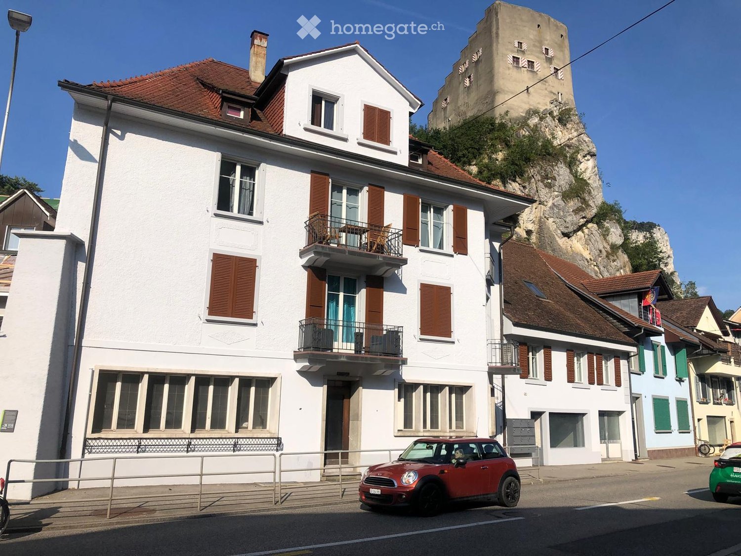 white building with brown roof, balconies, street view, red car parked in front