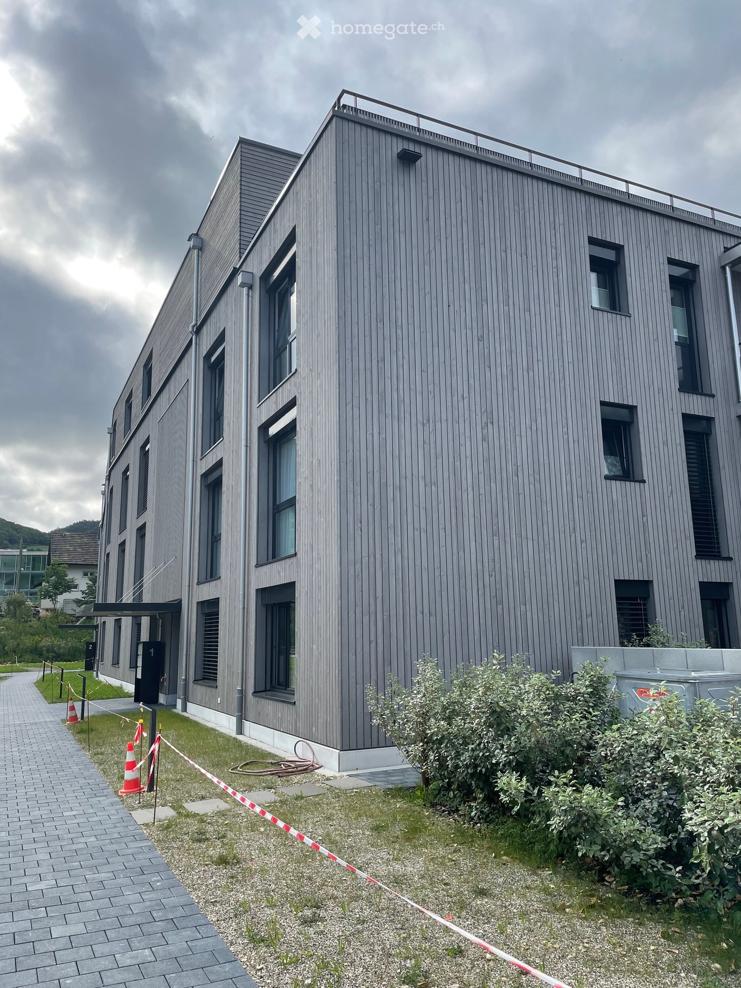 3 story building, gray exterior, multiple windows, entrance with awning, fenced area, pathway, bushes, construction site