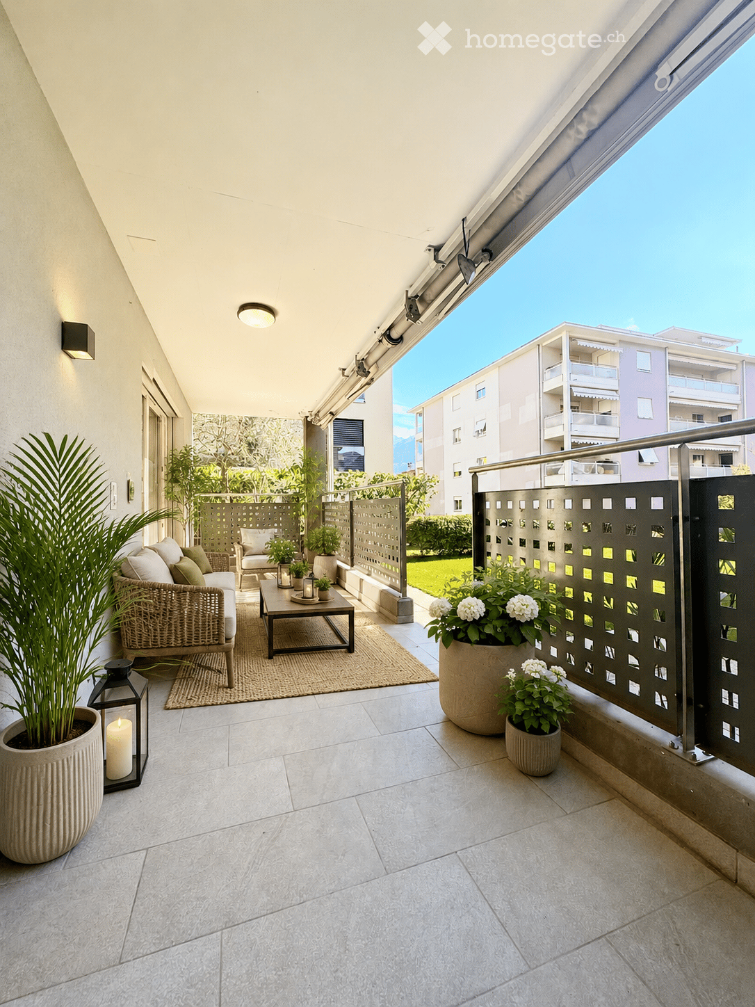 Balcony with modern furniture, including a sofa, coffee table, and lantern, surrounded by potted plants and a view of the city.