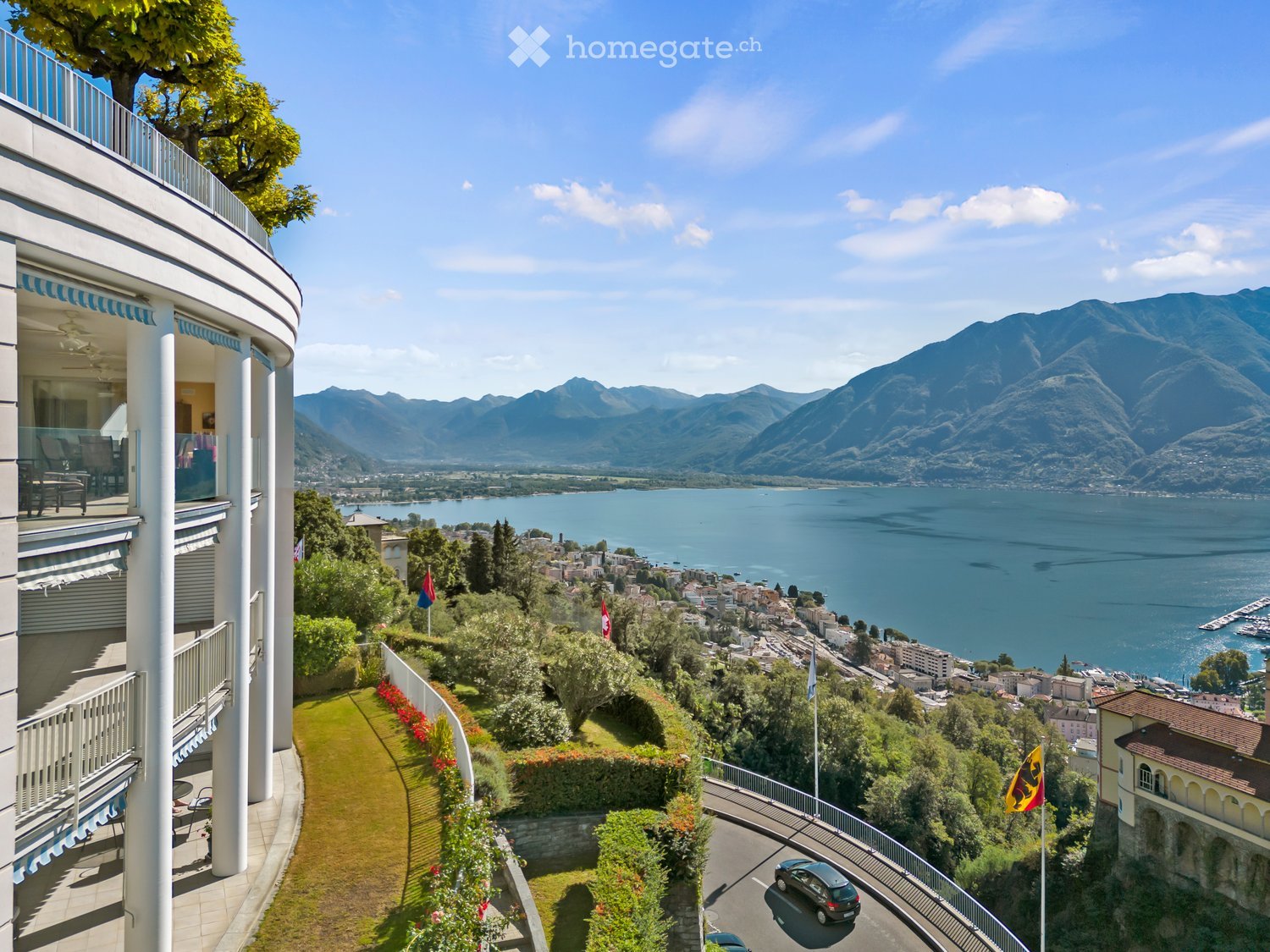 Mountainside house, balcony with railings, overlooking mountains and lake, fenced lawn, winding road, distant village