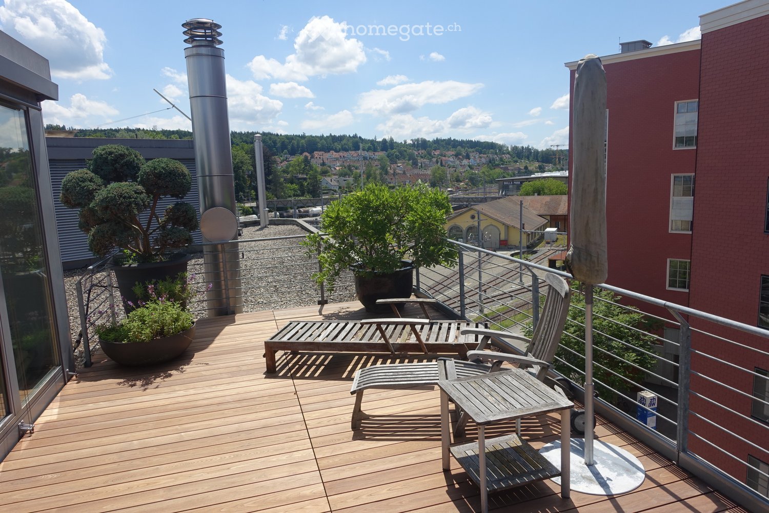 Wooden deck, metal railing, lounge chair, small table, potted plants, metal pole, view of the city