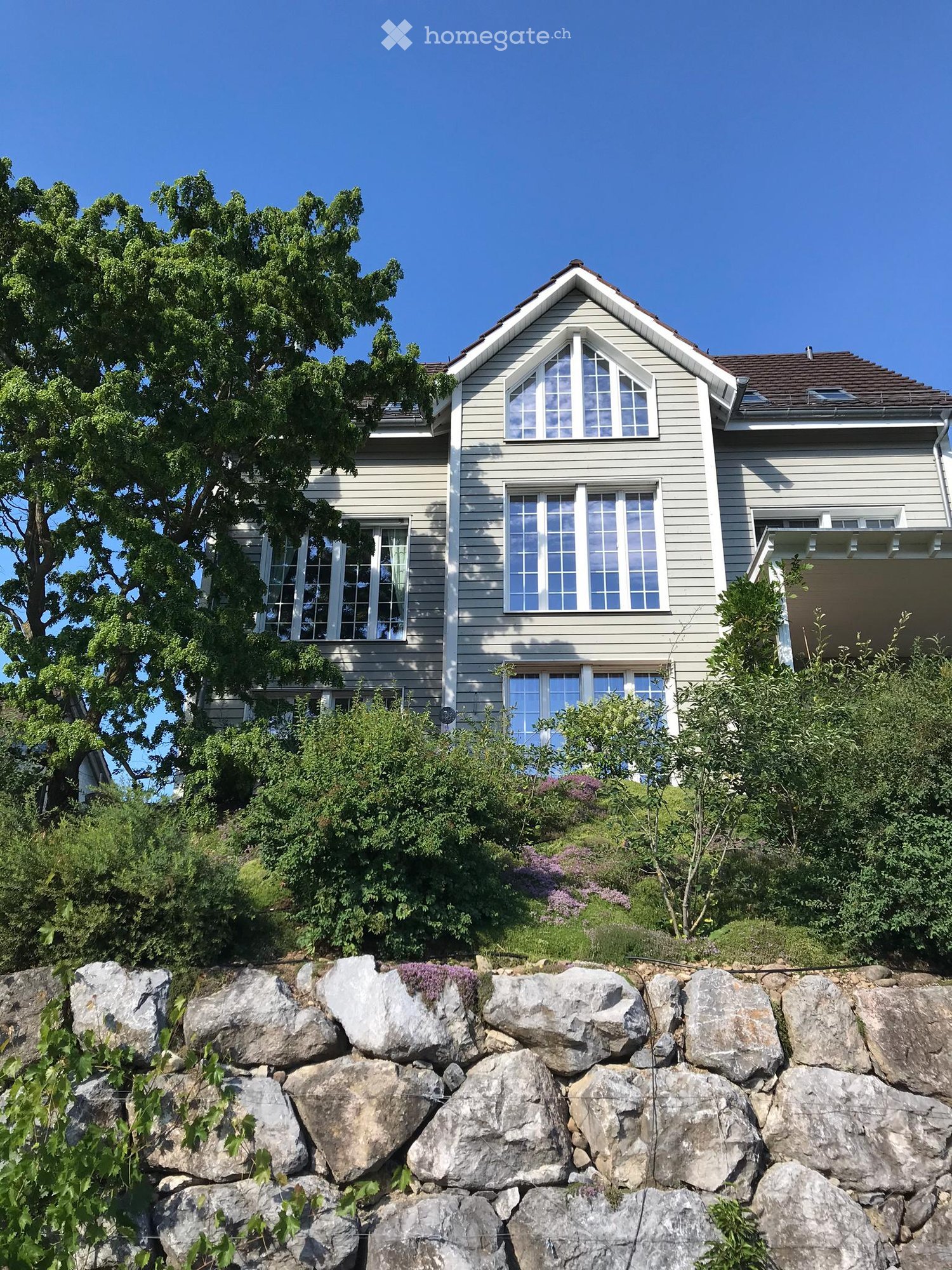 2 story house, gray exterior, large windows, brown roof, stone wall, garden, tree