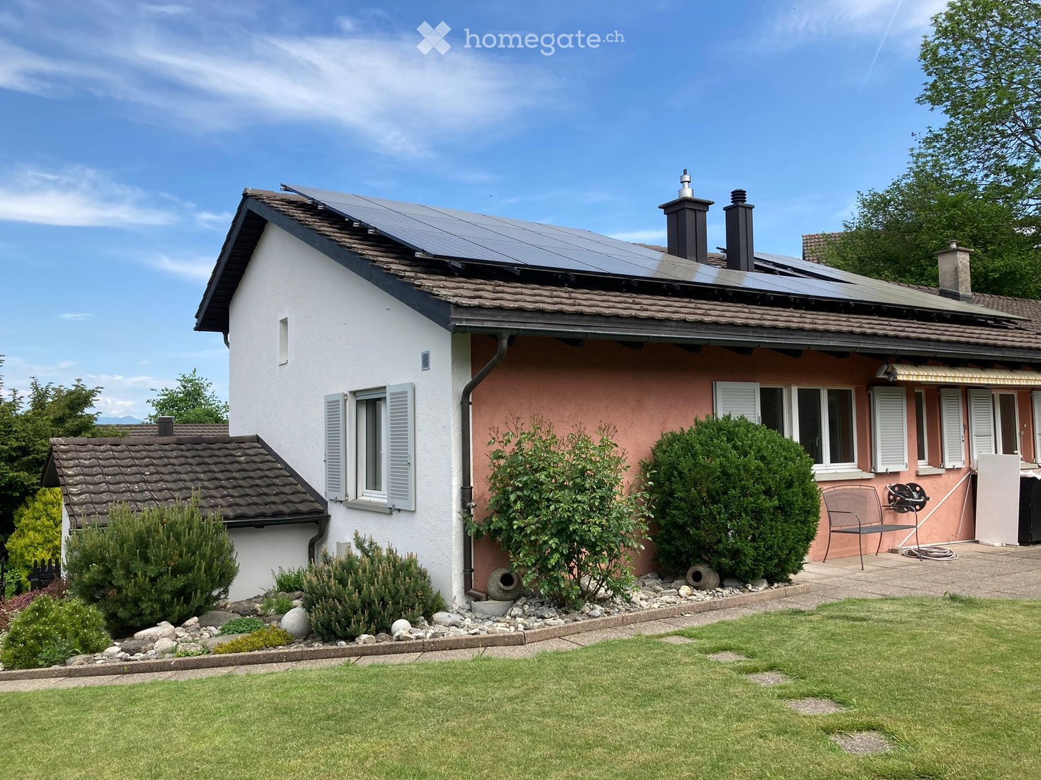 Detached house, solar panels on the roof, pink and white exterior, well-maintained garden, green lawn, shrubs, and plants, driveway, trees in the background