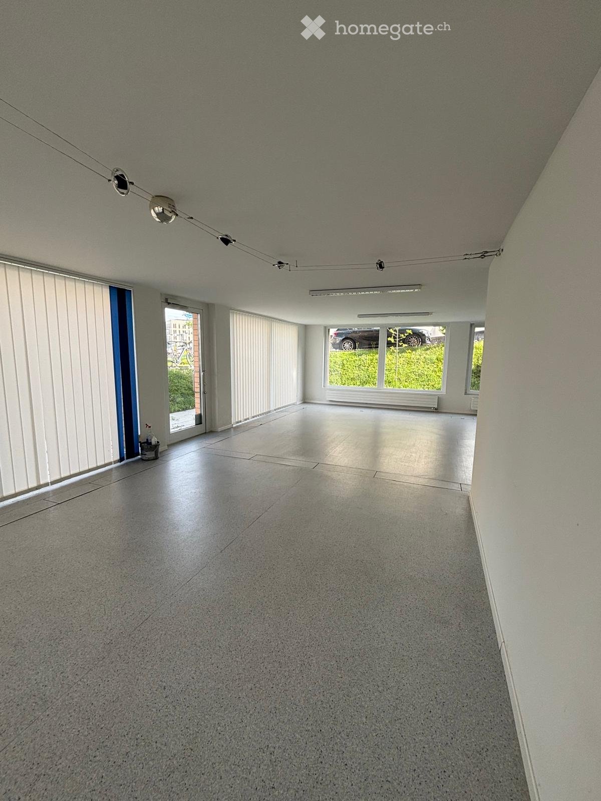 empty room, white walls, gray floor, large windows, white blinds, ceiling lights, door, parking area outside