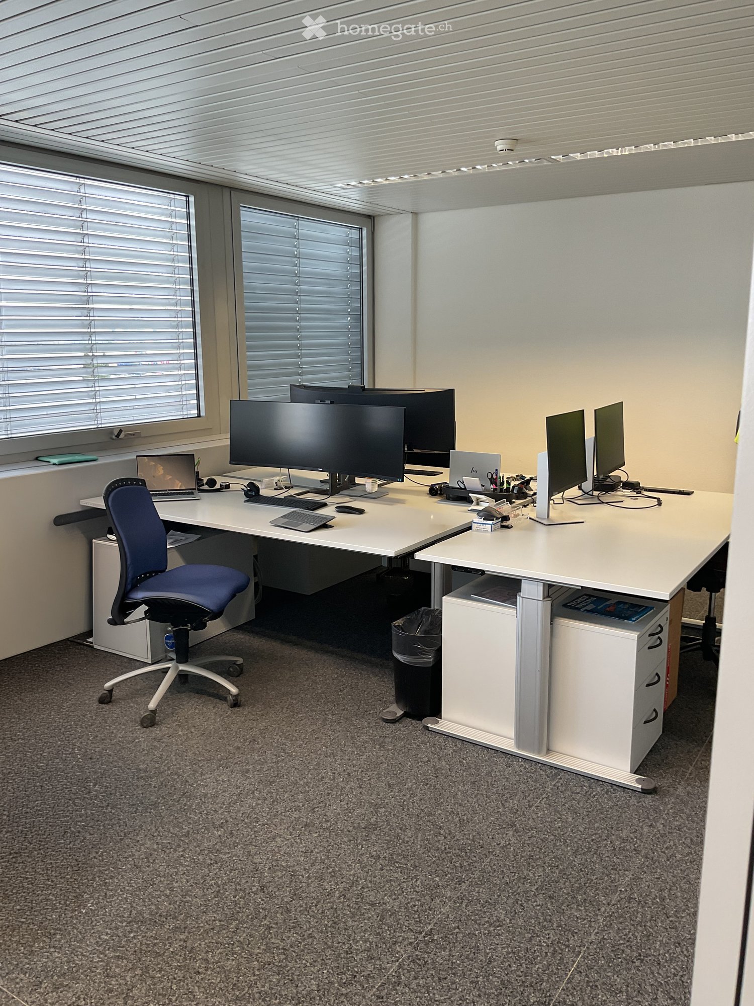 Two desks with monitors, chairs, cabinets, trash bin, windows with blinds